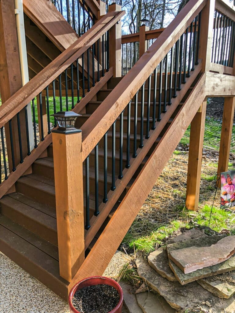 Wooden staircase with black railings, leading to a deck, surrounded by greenery and a potted plant, showcasing outdoor structure solutions by Home Deck and Fence Co.