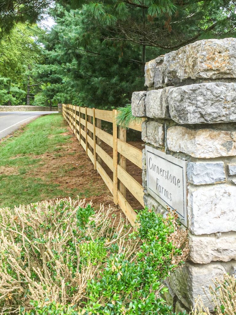Wooden fence along a road, bordered by lush greenery, featuring a stone pillar with a sign reading "Cornerstone Farms," highlighting quality fencing solutions for enhancing outdoor spaces.