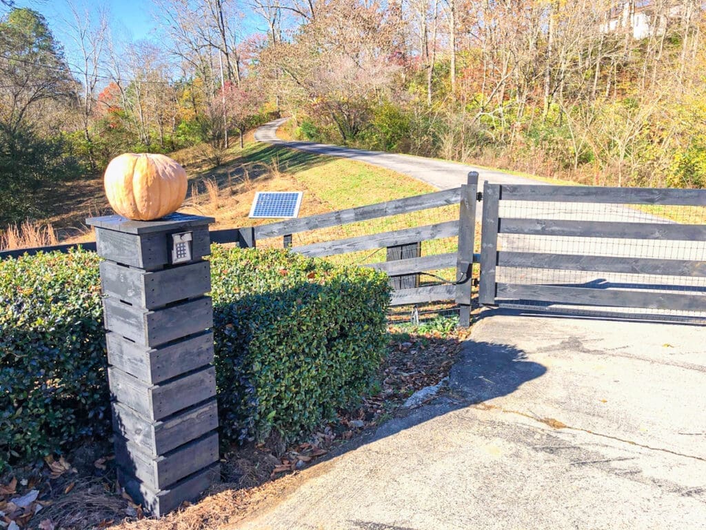 Black wooden gate with a pumpkin atop a post, surrounded by greenery, leading to a gravel driveway in a rural setting, reflecting home and fence aesthetics in Middle Tennessee.