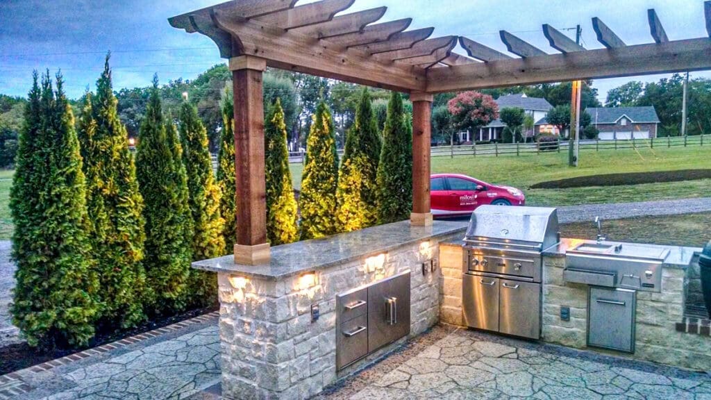 Outdoor kitchen with stainless steel grill and stone countertop, surrounded by tall green shrubs, showcasing custom outdoor construction by Home Deck & Fence Co. in Middle Tennessee.