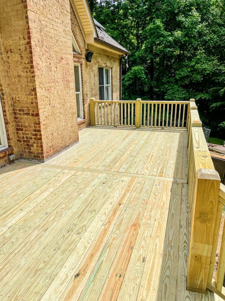 Newly constructed wooden deck with railings adjacent to a brick house, surrounded by lush greenery, showcasing outdoor living space enhancements in Middle Tennessee.