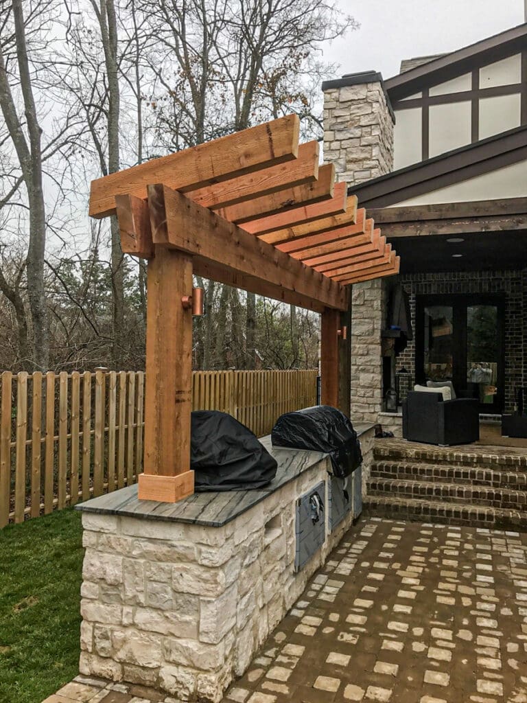 Custom wooden pergola over stone outdoor kitchen, featuring a brick patio and adjacent wooden fence, showcasing outdoor living space by Home Deck & Fence Co. in Dickson County, Tennessee.