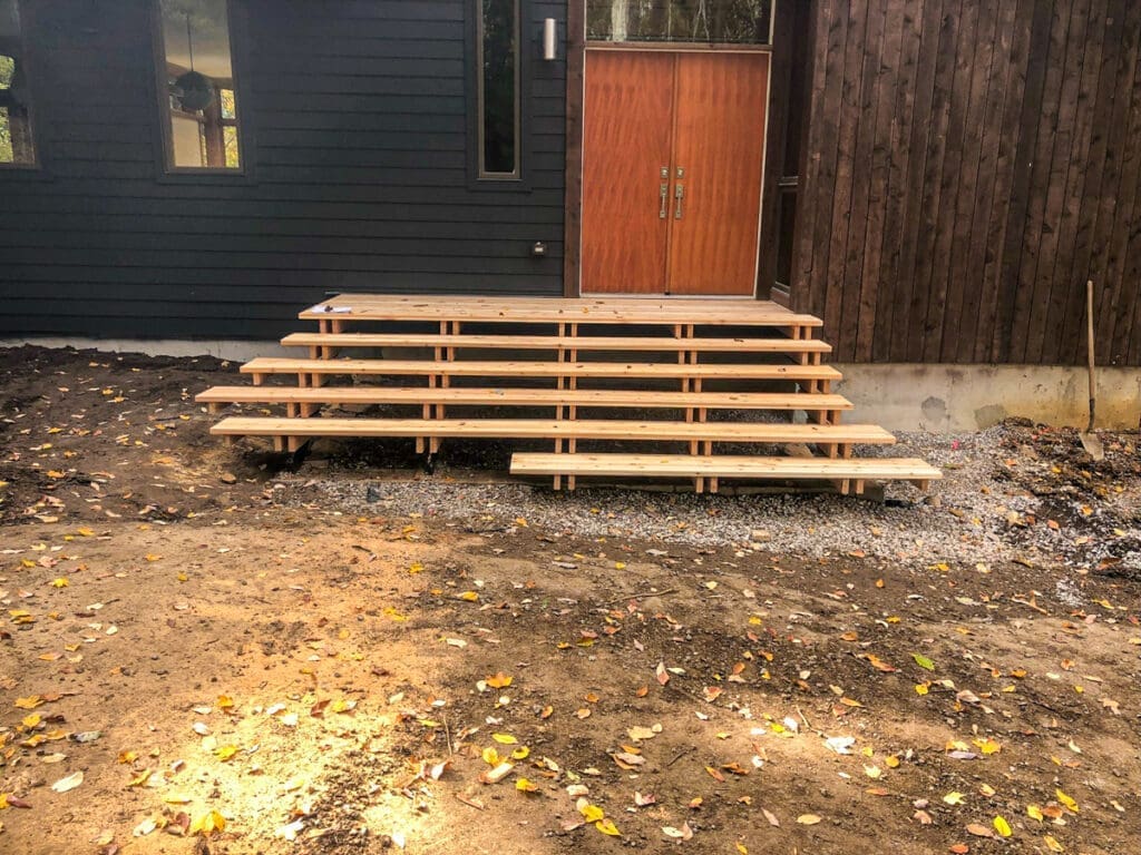 Wooden steps leading to a modern home entrance, featuring a double door, surrounded by gravel and fallen leaves, highlighting outdoor construction and design.