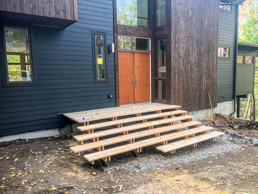 Wooden deck steps leading to a modern home entrance with large wooden doors, surrounded by a natural landscape in Dickson County, Tennessee, showcasing outdoor structure solutions by Home Deck and Fence Co.