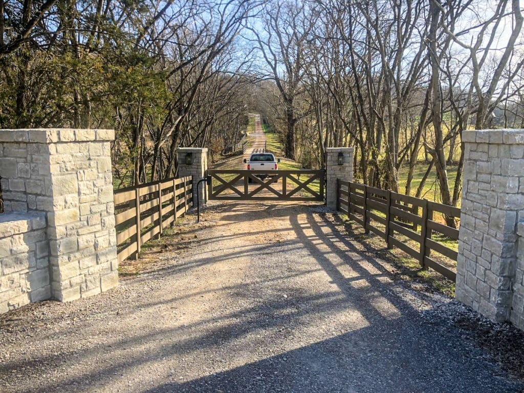 Stone and wooden gate entrance with a gravel driveway leading to a scenic pathway, surrounded by trees, representing quality fencing solutions by Home Deck & Fence Co.