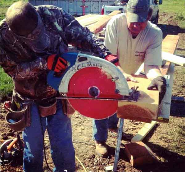 Two men collaborating on a woodworking project, one using a circular saw to cut a wooden plank, showcasing skilled craftsmanship in outdoor construction.