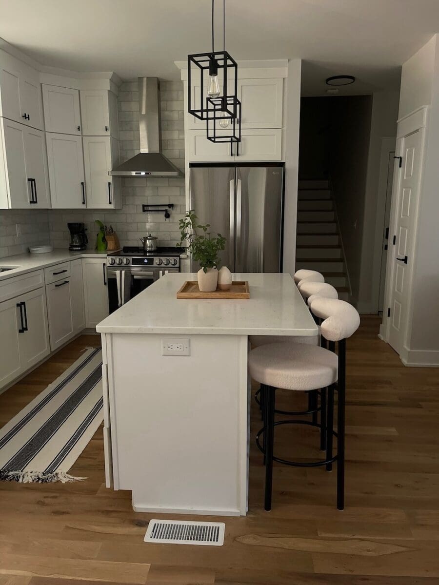 Modern kitchen featuring white cabinetry, quartz countertops, black pendant lighting, and a large center island with bar seating, complemented by warm hardwood floors and stylish decor.