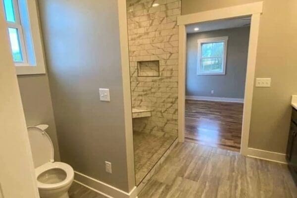 Modern bathroom interior featuring a walk-in shower with tiled walls, a toilet, and a doorway leading to a room with hardwood flooring, showcasing HDF Builders' custom home design.