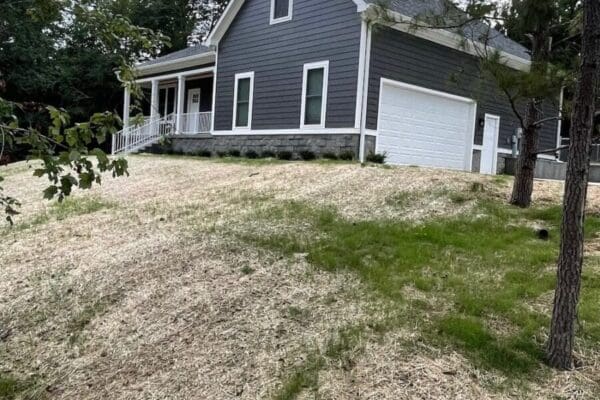 Newly constructed gray house with white trim, covered front porch, and two-car garage, situated on a sloped lot with fresh landscaping in a wooded area.