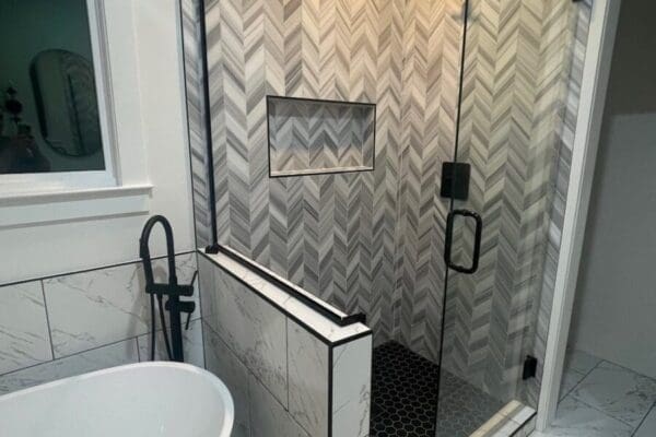 Modern bathroom featuring a sleek glass shower with herringbone tile pattern, black accents, and a freestanding tub, showcasing custom design elements for a luxurious feel.