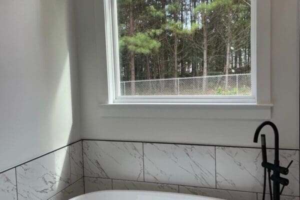 Modern bathroom featuring a freestanding tub, elegant marble tile, and a large window with wooded views, showcasing a serene and stylish design.