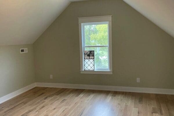 Vaulted bonus room with natural light from a window, showcasing fresh paint and hardwood flooring, ideal for guests or work in a custom-built home.