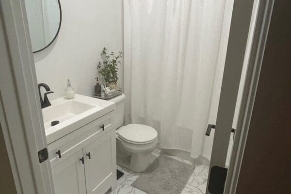 Modern bathroom with white cabinetry, round mirror, and shower curtain, featuring a clean design and decorative plant, reflecting stylish guest bath elements.