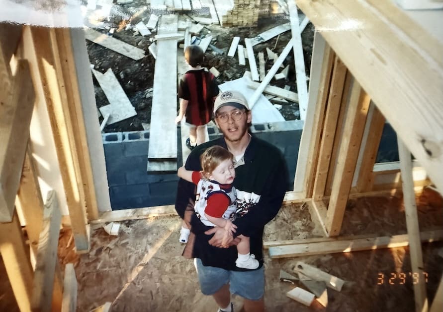 Father holding child in a construction site, with wooden framing and debris in the background, symbolizing family involvement in building and craftsmanship.